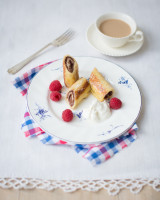 Cup of coffee next to a plate with rolled-up chocolate toasts, next to raspberries and yoghurt