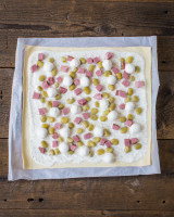 Rolled-out dough on baking paper, topped with white sauce, gardenbeans, mini mozzarella balls and pork cubes