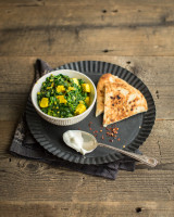 Bowl of Paneer cheese and spinach on a black plate with naan bread and a spoon with yoghurt